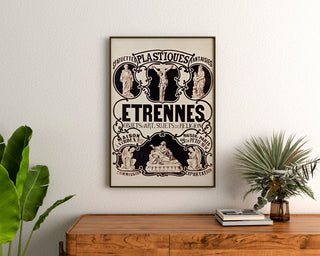Vintage reklameplakat Verdun, statuetter og nytårsgaver, indrammet over en trækommode mellem planter.