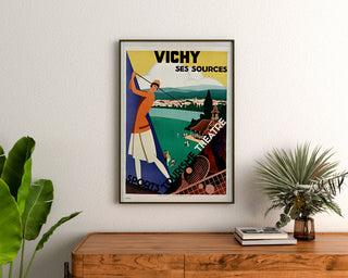 Vintage turistplakat – Vichy, kilder og turisme, indrammet over en trækommode mellem planter.