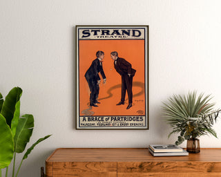 Vintageplakat A Brace of Partridges, Strand Theatre, indrammet over en trækommode mellem planter.